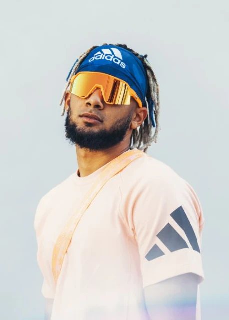 Portrait of a male adult wearing a blue Adidas headband, orange-tinted sunglasses, and a salmon-colored t-shirt, set against a light background.