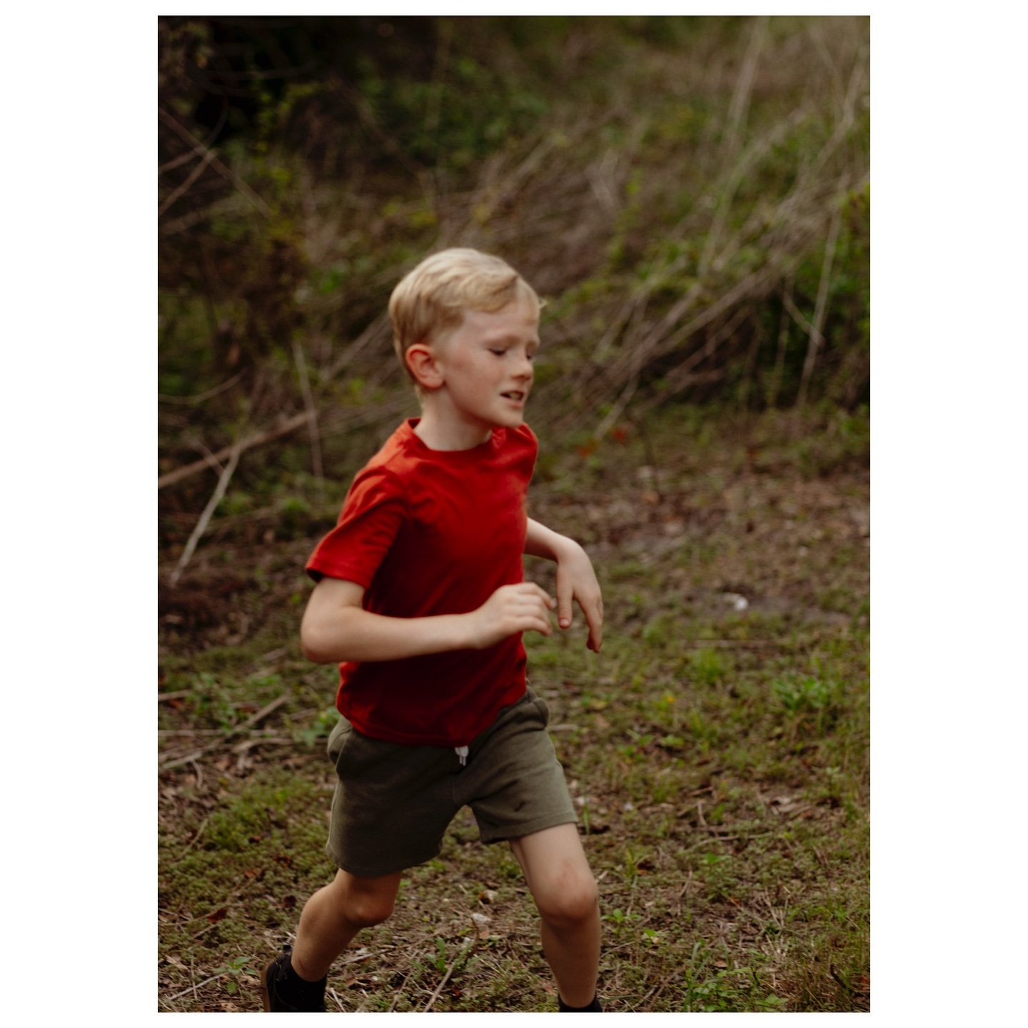 A young blond boy wearing a red shirt runs through a wooded area on a cloudy day, his face expressing energy and determination.