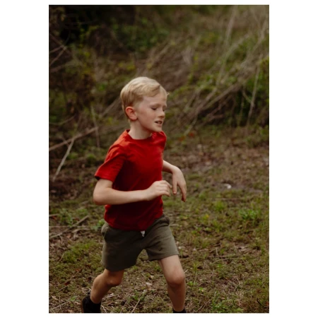 A young blond boy wearing a red shirt runs through a wooded area on a cloudy day, his face expressing energy and determination.