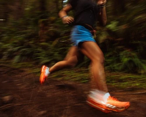 An athlete is motion-blurred running on a trail, showing only the lower portion of their body in motion. The background is dense forest.