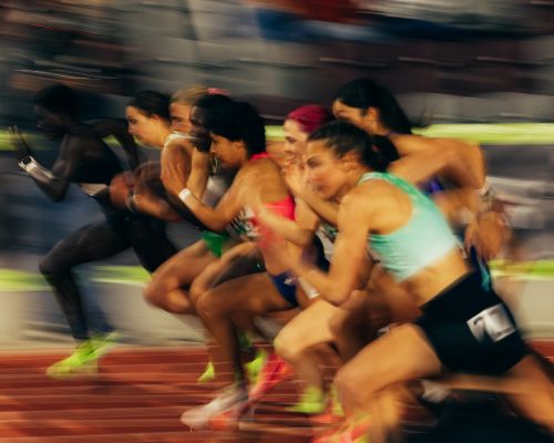 Motion blur photo of diverse group of women athletes running at a track competition, wearing sportswear and bright running shoes.