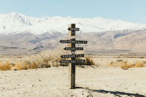 Wooden signpost with multiple directional signs points to hiking trails in a desert landscape with snow-capped mountains in the background.