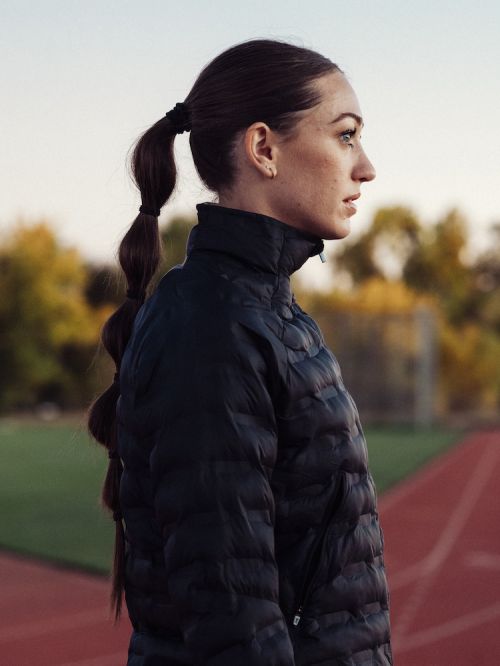 Profile shot of a female athlete with a high ponytail, wearing a black puffer jacket, standing on a red running track outside.