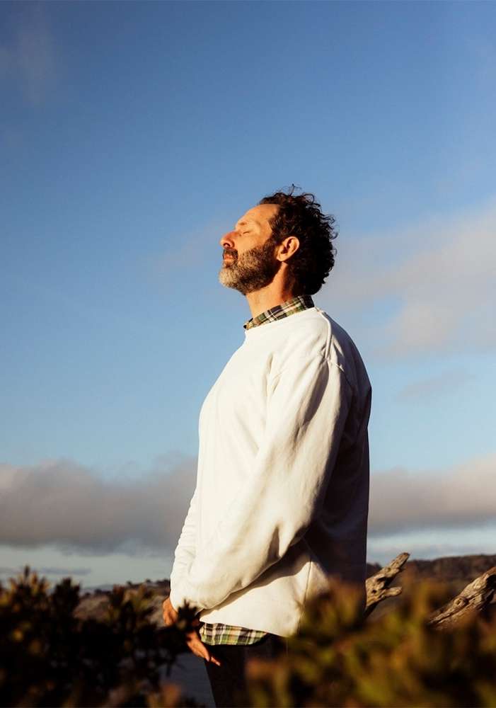 Adult man with eyes closed standing outdoors wearing a white sweater, a patterned shirt and blue sky in the background.