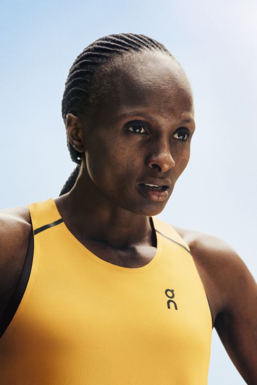 Close-up of a serious African-American adult female athlete wearing a yellow and black tank top with braided hair against a blue sky.