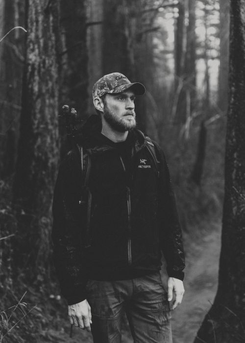 Monochrome portrait of a bearded man in a camo cap and jacket standing on a trail in a dark, wooded forest.