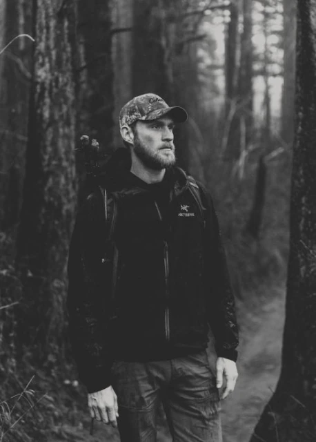 Monochrome portrait of a bearded man in a camo cap and jacket standing on a trail in a dark, wooded forest.
