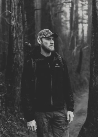 Monochrome portrait of a bearded man in a camo cap and jacket standing on a trail in a dark, wooded forest.