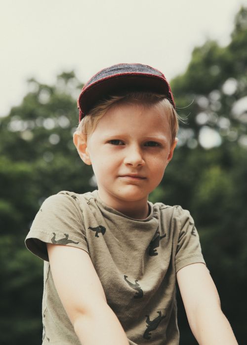 Portrait of a young boy with blond hair wearing a baseball cap outdoors, looking directly at the camera against a blurred tree background.