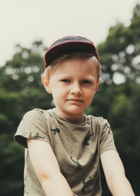 Portrait of a young boy with blond hair wearing a baseball cap outdoors, looking directly at the camera against a blurred tree background.