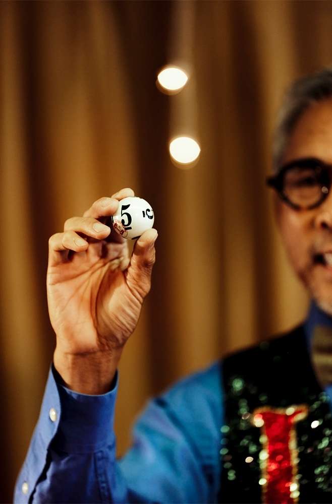 Adult male in a blue shirt and glasses holds up a lottery ball numbered 5 in a close-up studio shot with a curtain backdrop.