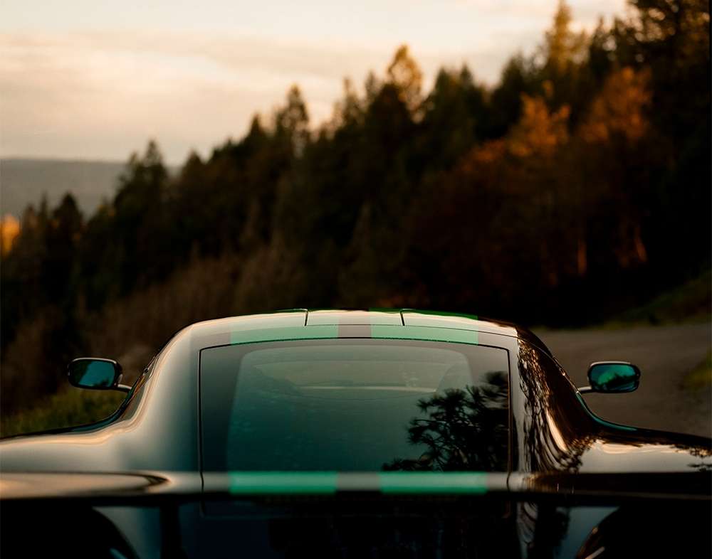 Rear view of a sleek black sports car with green stripes parked on a road, with a scenic backdrop of sunset and mountain forest.