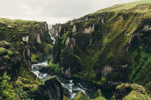 Scenic view of a canyon with a river flowing through it, surrounded by moss-covered cliffs and lush greenery under an overcast sky.