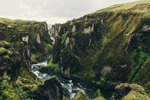 Scenic view of a canyon with a river flowing through it, surrounded by moss-covered cliffs and lush greenery under an overcast sky.
