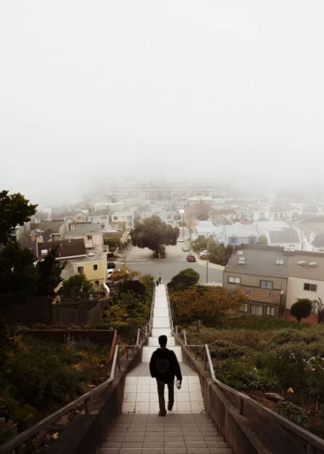 Person walks down concrete stairs in an urban setting with fog over a city and tree, photographed from an eye-level angle.