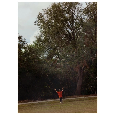 Child in an orange shirt with arms raised running on a grassy field toward a large tree line under an overcast sky.