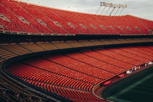 High-angle view of an empty American football stadium with red and yellow seating under an overcast sky, showcasing its vast structure.