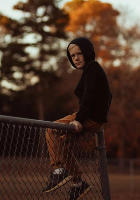 Young person in a hoodie sits on a chain-link fence in a park with fall foliage.  Portrait captures the thoughtful moment of the person in an outdoor setting.