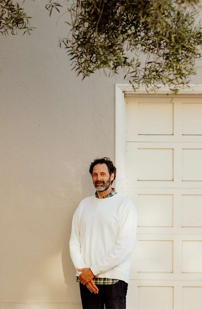 Man standing against an off-white wall next to a white panel door beneath leafy olive branches.