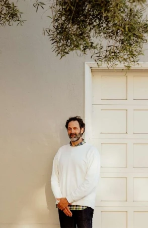 Man standing against an off-white wall next to a white panel door beneath leafy olive branches.