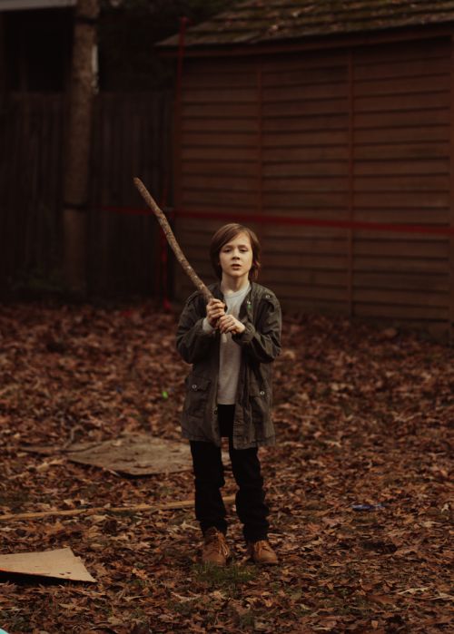 Young person in jacket stands in yard holding a stick. Leaves are scattered on the ground, a building sits in the background.