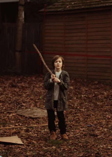 Young person in jacket stands in yard holding a stick. Leaves are scattered on the ground, a building sits in the background.