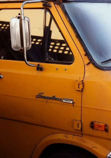 Close-up of a vintage yellow Ford Econoline 100 van featuring a side mirror, front window, door, and the Econoline 100 emblem.