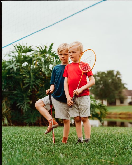 Two young boys stand on a green lawn; one holds a badminton racket, and the other rests his foot on a second racket in a yard setting.