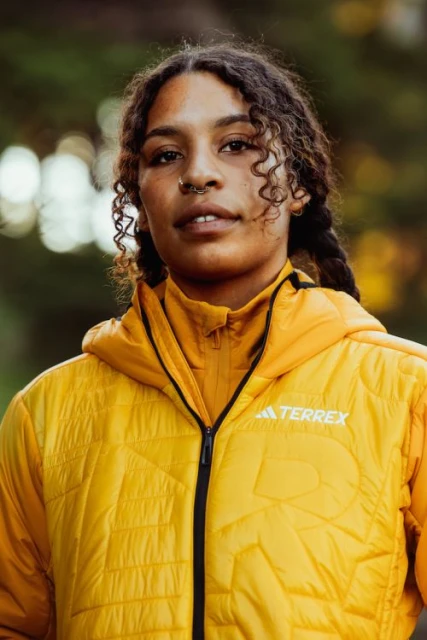 Close-up of an adult female with curly dark hair and a nose ring, wearing a yellow hooded jacket with the Adidas Terrex logo, outdoors.
