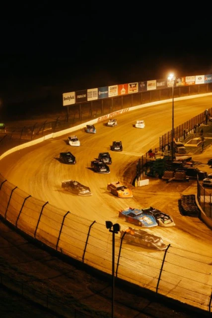 Nighttime dirt track racing event featuring a group of race cars speeding around a banked oval track under bright stadium lights.