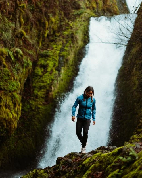 Woman in blue jacket hikes on mossy rocks with a powerful waterfall cascading in the background through lush green foliage.