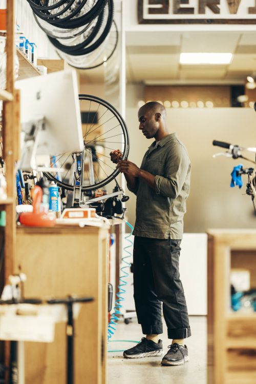 Adult male repairing a bike tire inside a bike shop. Bike wheels are hanging on the wall. The man is wearing a green shirt.