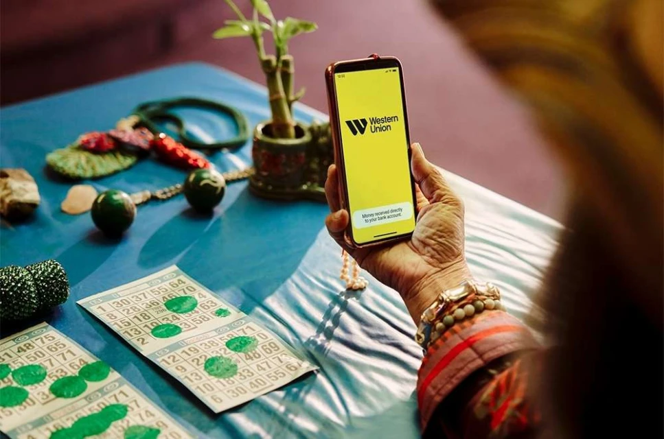 An older adult holds a phone displaying the Western Union app, with bingo cards and other objects on a blue surface.