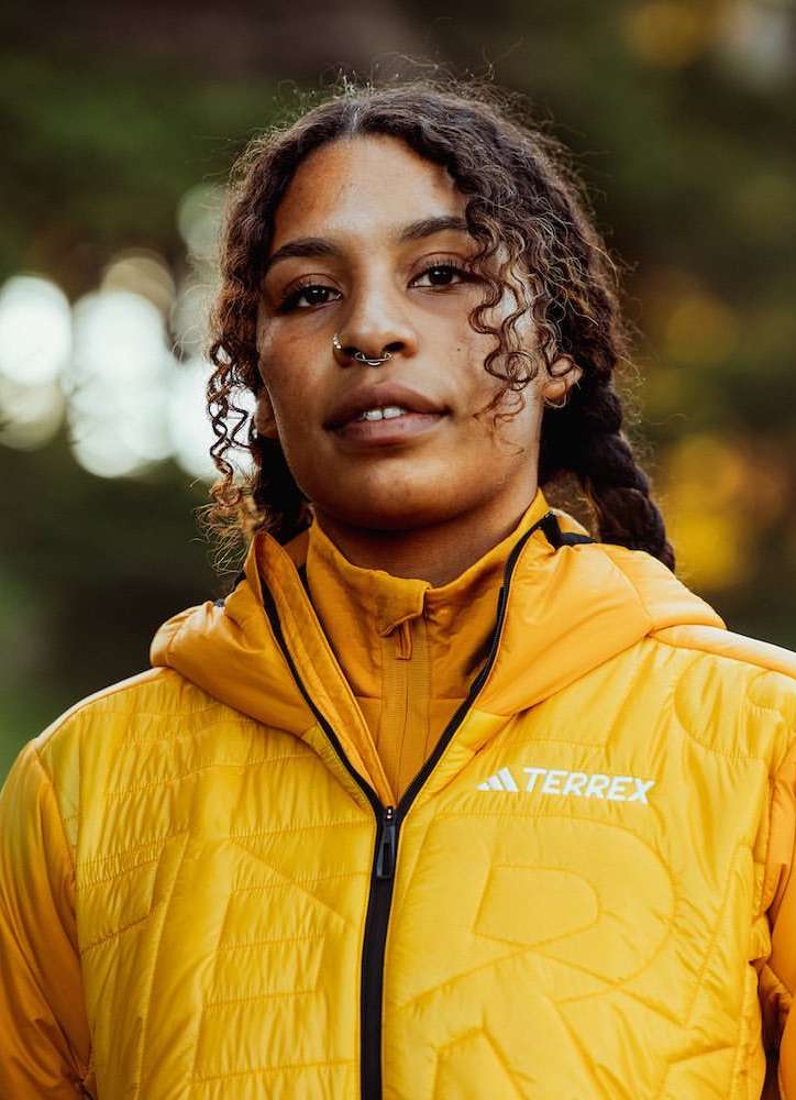 Portrait of a curly-haired woman wearing a yellow hooded jacket in a wooded area, looking directly at the camera with a neutral expression.