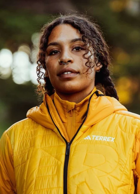 Portrait of a curly-haired woman wearing a yellow hooded jacket in a wooded area, looking directly at the camera with a neutral expression.