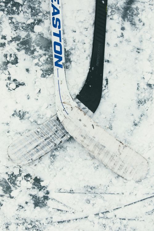 Two hockey sticks are crossed on a snowy surface outdoors. An Easton brand stick is visible, lying on top of a black hockey stick.