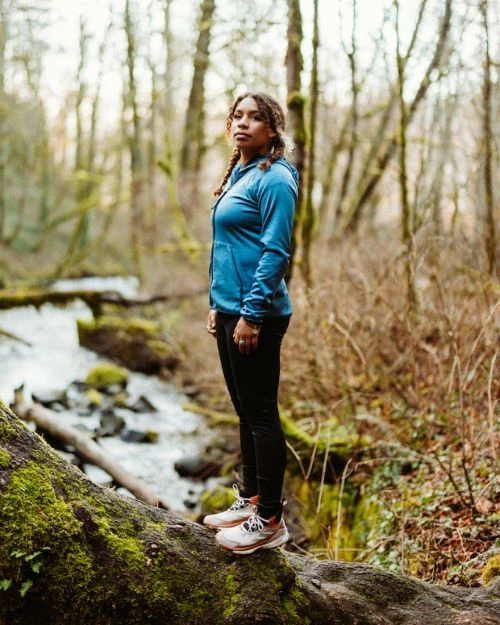 A woman in outdoor sportswear stands on a mossy log near a creek in a lush forest. She's wearing a blue jacket and black leggings.