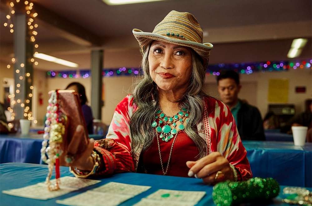 Adult woman in hat holds beaded jewelry with cell phone at bingo table in decorated hall with warm lighting and another adult in background.