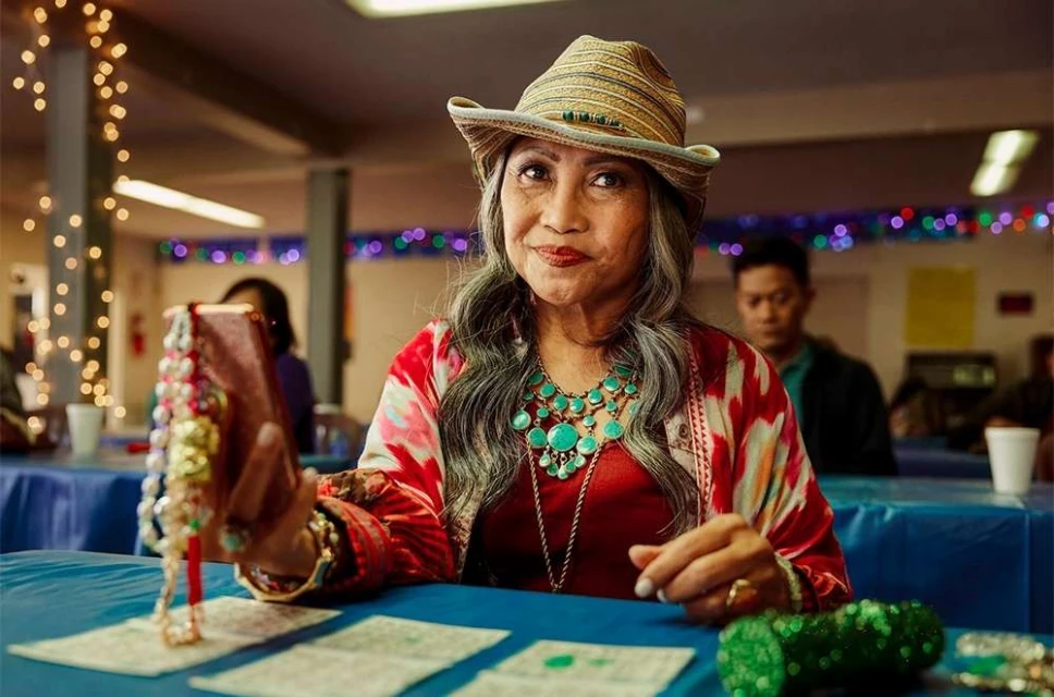 Adult woman in hat holds beaded jewelry with cell phone at bingo table in decorated hall with warm lighting and another adult in background.