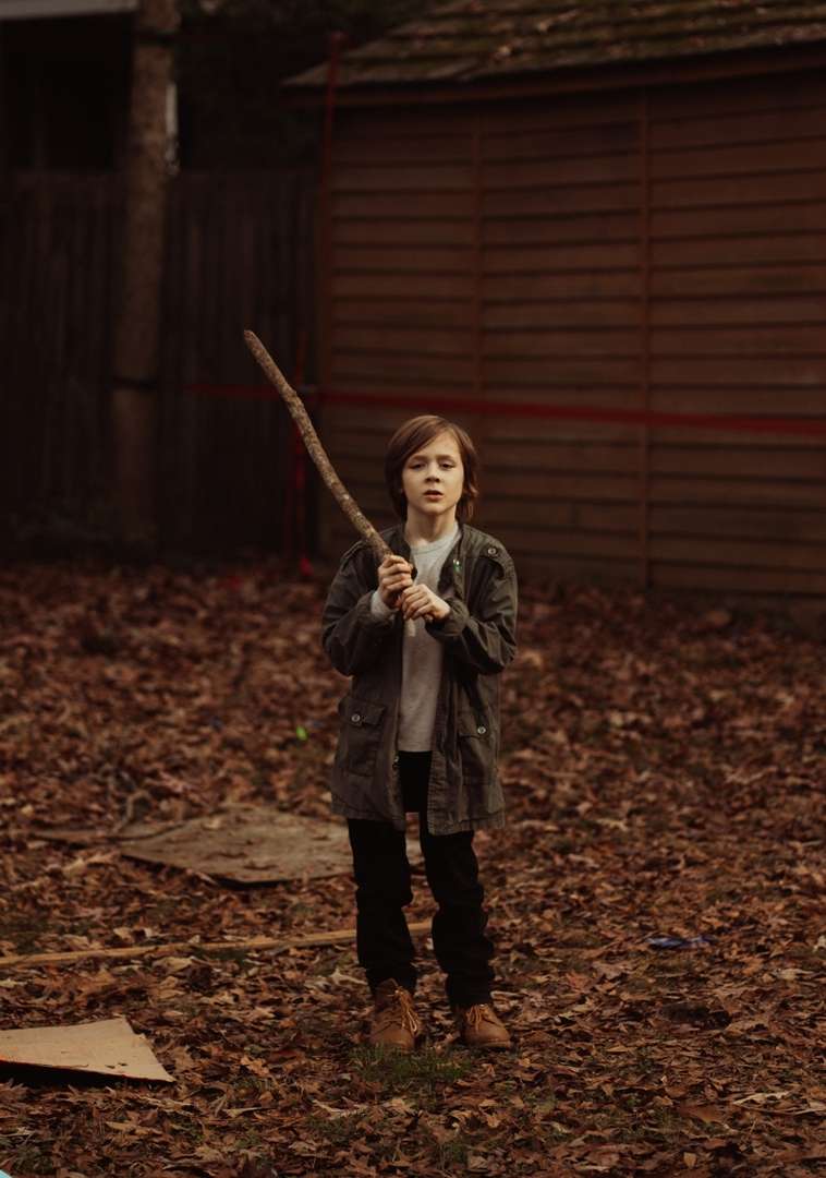 A young boy stands outdoors in autumn leaves, holding a large stick, wearing a jacket, pants, and tan boots, with trees and a shed in the background.