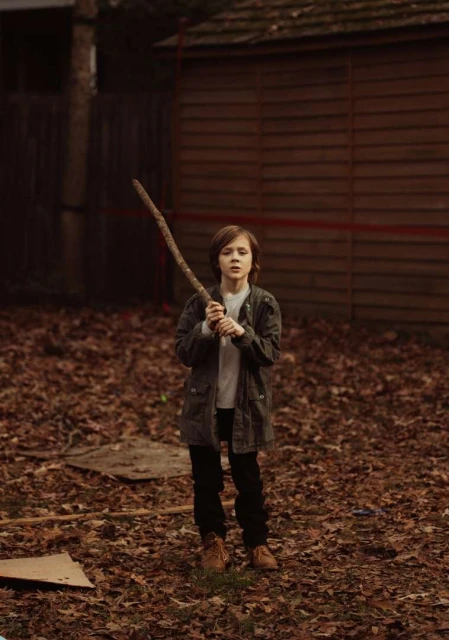 A young boy stands outdoors in autumn leaves, holding a large stick, wearing a jacket, pants, and tan boots, with trees and a shed in the background.