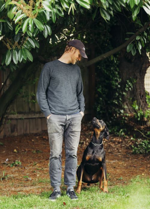 Adult man standing in a yard with a young black and tan Doberman dog, looking downward in daylight, underneath a tree canopy.