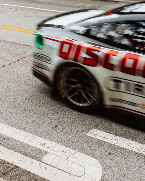 Blurred motion shot of a race car on asphalt racetrack with red Discount Tire livery and yellow lane divider lines on the road surface.