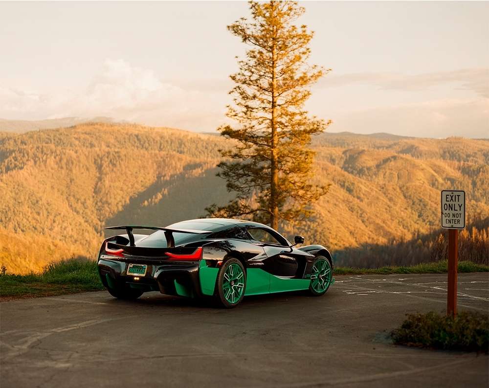 Black and green Rimac Nevera parked on an overlook with a view of forested mountains and a tall pine tree during golden hour.