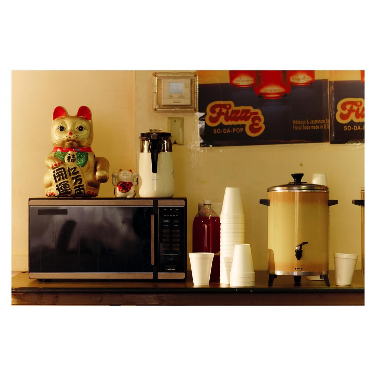 Indoor shot of a beverage station with a drink dispenser, cups, beverage bottles, microwave, and lucky cat figurine on a shelf.