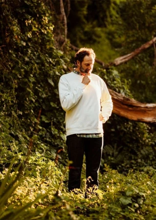 Man standing outdoors in a white sweatshirt and dark trousers amidst dense green foliage and trees.