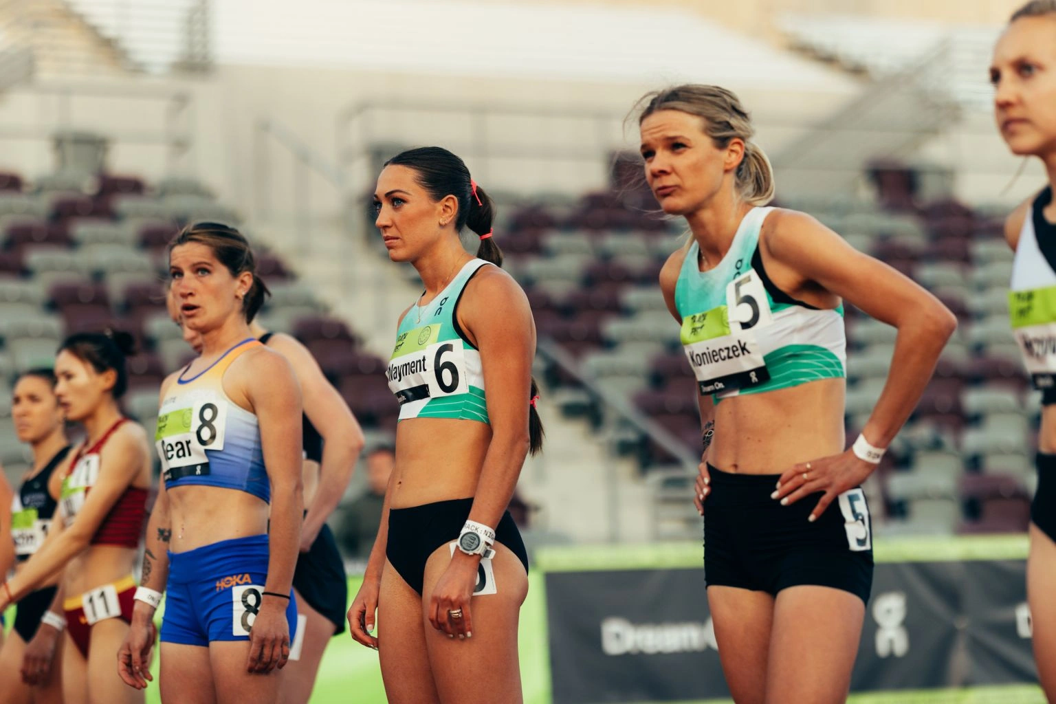 Group of female athletes lined up on a running track wearing athletic crop tops and race bibs, photographed by Kennett Mohrman.