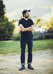 Man with beard wearing dark t-shirt, baseball cap, and cuffed jeans standing outdoors with arms crossed.