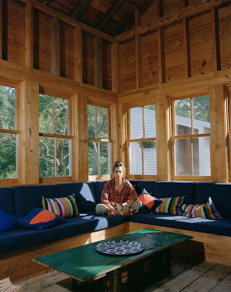Indoor portrait of a woman sitting cross-legged on a blue cushioned bench seat with colorful pillows, with a green table in the foreground.