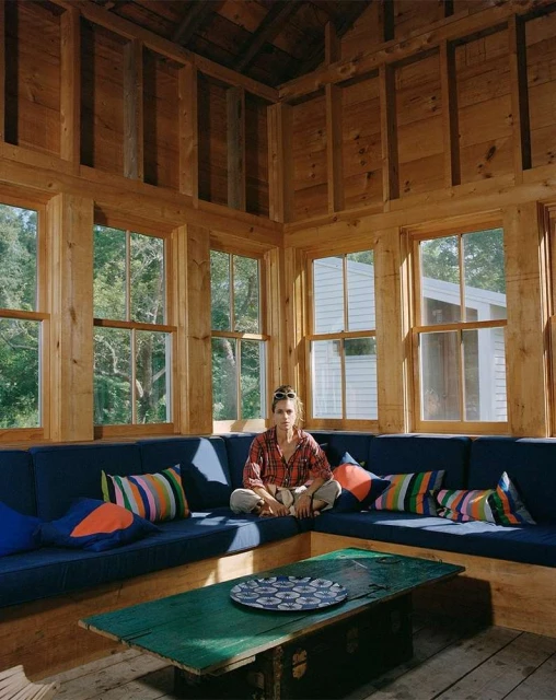 Indoor portrait of a woman sitting cross-legged on a blue cushioned bench seat with colorful pillows, with a green table in the foreground.